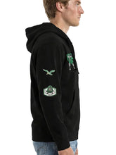 A man shown in profile wearing a black pullover hoodie with green and white graphic patches on the chest and sleeves, paired with blue jeans, against a light gray studio background.
