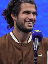 A smiling man with dark curly hair and a beard, wearing a brown mesh baseball-style jacket with white trim, standing in front of a blue backdrop and a purple microphone that says US Open.