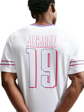 Back view of a white short-sleeve jersey with 'ALCARAZ' and the number '19' outlined in red and blue, worn by a model against a light gray background.