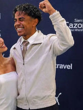 A smiling man in a white zip-up jacket over a white shirt and tie raises his fist in front of a dark blue branded backdrop. Part of a woman in a white top is visible at the left edge.