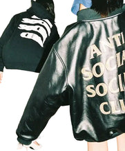 A close-up of a person wearing a black leather jacket with large gold lettering on the back, standing in a dimly lit indoor setting. Another person in a black hoodie is partially visible in the background.