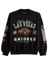 A black crewneck sweatshirt featuring the Las Vegas Raiders logo and name in white and silver, with a central brown football graphic, shown on a light gray background.
