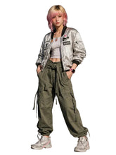 A model with pink hair stands against a concrete wall wearing a shiny silver bomber jacket, olive green cargo pants, a white crop top, and white sneakers.