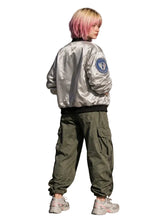 A person with pink-tinted hair stands in profile wearing a shiny silver bomber jacket with a blue circular patch on the sleeve, olive green cargo pants, and white sneakers, set against a dark concrete wall.