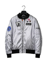 A silver satin bomber jacket with black ribbed trim, hanging on a black hanger against a dark background under a spotlight. The jacket features multiple embroidered patches, including a 'VISITOR' patch and a blue circular patch on the sleeve.