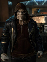 A person wearing a dark hooded jacket and matching dark top, standing in a dim indoor environment with blurred equipment or screens in the background.