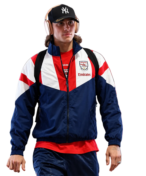 A man wearing an Arsenal tracksuit jacket and matching pants walks indoors while wearing a black New York Yankees cap and over-ear headphones. Another person is partially visible behind him.