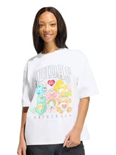 A woman wearing a white Adidas Originals graphic t-shirt featuring Care Bears characters, set against a solid light gray background.