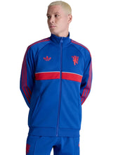 A man wearing a blue and red Manchester United track jacket by Adidas, featuring the club's crest on the chest, a red horizontal stripe, and red three-stripes on the sleeves, set against a plain light gray background.