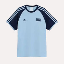 A light blue and navy blue raglan t-shirt featuring the Adidas Trefoil logo on the left chest and a 'oasis' brand patch on the right chest, with three white stripes on the sleeves, shown against a plain off-white background.
