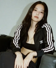 A woman wearing a black Adidas track jacket with white stripes and a black sports bra, sitting on a dark leather couch against a light gray wall.