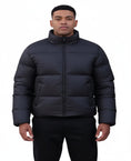 A man in a black puffer jacket stands behind another person wearing a beige coat with a dark fur-trimmed collar, against a plain white background.