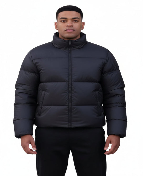 A man in a black puffer jacket stands behind another person wearing a beige coat with a dark fur-trimmed collar, against a plain white background.