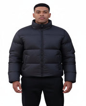 A man in a black puffer jacket stands behind another person wearing a beige coat with a dark fur-trimmed collar, against a plain white background.
