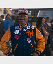A man wearing a New York Mets varsity jacket with orange sleeves, a navy blue body, and multiple patches, including a 'NEW YORK' baseball cap, standing outdoors at night.
