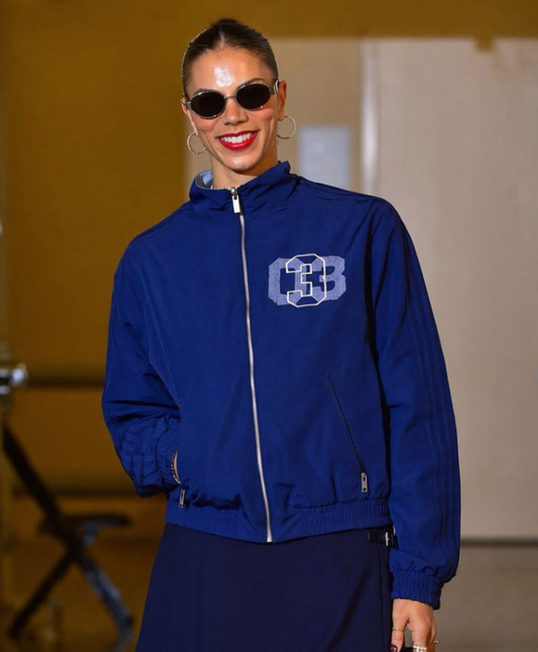 A woman wearing a royal blue zip-up jacket with a white and blue '3B' monogram on the chest, paired with a dark skirt, photographed on a runway or stage.