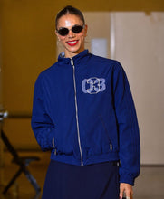 A woman wearing a royal blue zip-up jacket with a white and blue '3B' monogram on the chest, paired with a dark skirt, photographed on a runway or stage.