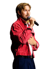A man with blond hair and a beard stands on a stage holding a microphone, wearing a red jacket over a white shirt and dark trousers. Comic-Con International logos are visible in the background.