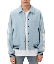A man wearing a light blue denim varsity-style jacket with cream-colored distressed graphics on the sleeves, black and navy striped ribbed trim, and a white t-shirt underneath, set against a plain white background.