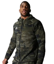 A man wearing a camouflage zip-up hoodie with a white circular logo on the left chest, standing in an indoor industrial setting.