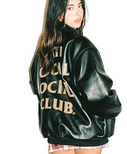 A woman wearing a black leather bomber jacket with gold 'THE SOCIAL CLUB' text on the back, photographed against a plain white background.