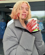 A woman with short blonde hair wearing a gray puffer jacket, holding a white coffee cup covered in fake red blood splatter, with fake blood on her face and hand.