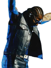 A man wearing a black leather vest and jeans performing on stage under dramatic blue lighting, with one arm raised high.