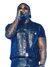 A tattooed performer on stage wearing a black leather vest over a dark blue short-sleeve shirt, with silver sunglasses and jewelry, holding a microphone against a bright blue background.
