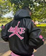 A person viewed from behind wearing a black leather hooded jacket with a large pink Jordan Wings logo on the back, standing outdoors in a park with trees and grass in the background.