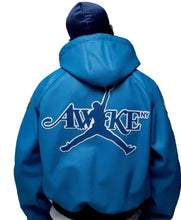 A person viewed from behind wearing a bright blue hooded jacket with a large white and blue 'AWAKE NY' graphic across the back, paired with a dark blue beanie, against a neutral gray studio background.