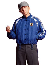 A man wearing a royal blue traditional Chinese-style jacket with white stripes on the sleeves, a gray cap, and dark trousers, standing against a solid red background.