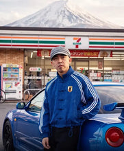 A man wearing a blue traditional-style jacket with white stripes and a gray cap stands next to a blue sports car in front of a 7-Eleven store and a snow-capped mountain.