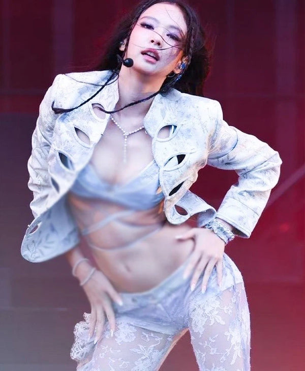 A woman posing on a stage in a light silver-gray lace and satin two-piece outfit with a matching cutout jacket, under red stage lighting.
