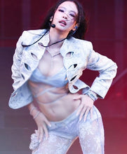 A woman posing on a stage in a light silver-gray lace and satin two-piece outfit with a matching cutout jacket, under red stage lighting.