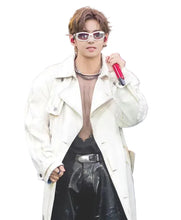 A male performer on stage wearing a long cream-colored trench coat over a sheer brown top and black pants, holding a red microphone against a dark background with blue stage lights.