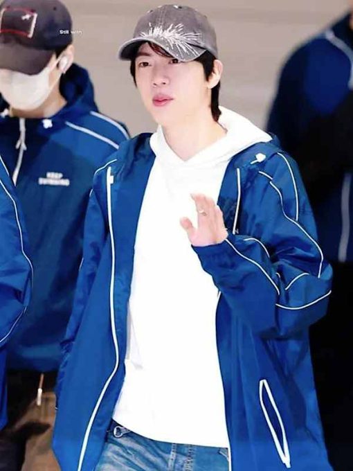 A man wearing a blue zip-up jacket with white piping over a white hoodie, blue jeans, and a gray patterned baseball cap, gesturing with his hand.