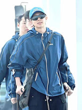 A man wearing a blue windbreaker jacket, matching blue cap, and black sunglasses, carrying a black shoulder bag while walking outdoors.