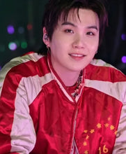 A close-up shot of a smiling young man wearing a shiny red and white satin jacket with yellow stars on the chest, set against a dark background with colorful bokeh lights.