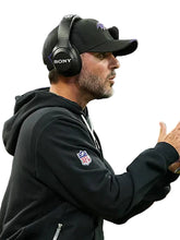 A man in a black NFL hoodie and black cap wearing a black Sony over-ear headset, shown in profile against a blurred stadium background.