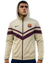 A man wearing a beige and purple FC Barcelona Nike zip-up hoodie jacket, shown from the waist up against a light gray background.