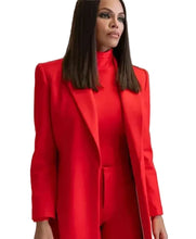 A woman wearing a bright red tailored blazer over a matching red turtleneck top and red pants, standing in a modern indoor hallway. A blurred man in a light gray suit is visible in the background.