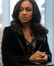 A woman with long, wavy black hair is seated indoors, wearing a black velvet blazer over a black lace top. Her hands are clasped together on a dark surface in front of her.