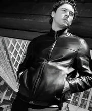 A black leather zip-up jacket worn by a man in a black-and-white photograph, posed against an urban architectural background.