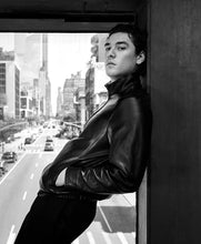 A black and white fashion photograph of a man wearing a black leather jacket and dark trousers, leaning against a dark pillar next to a window with a city street view.