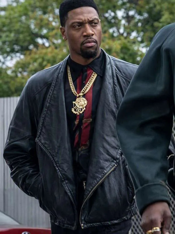 A man wearing a black leather jacket, a large gold chain necklace with a medallion pendant, and a red patterned shirt, standing outdoors.
