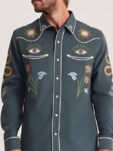A man wearing a dark slate blue long-sleeve button-up shirt with white piping and detailed embroidered motifs including suns, eyes, flowers, and leaves.
