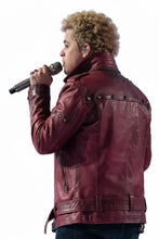A man with curly blonde hair singing into a microphone, wearing a dark red leather jacket with metal studs, with a band performing in a stage setting.