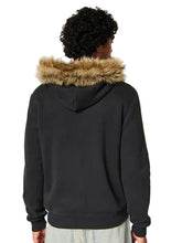 A person shown from the back wearing a black pullover hoodie with a tan faux-fur trimmed hood, against a plain light beige studio background.
