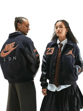 Two models wearing matching dark navy varsity-style jackets with orange Nike and Jordan logos, shown against a light gray studio background.