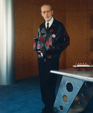 An older man with glasses, a gray beard, and a white shirt stands wearing a black leather bomber jacket covered in red and white patches, black trousers, and a black tie. He is posed in front of a textured brown wall next to a modern metallic table.
